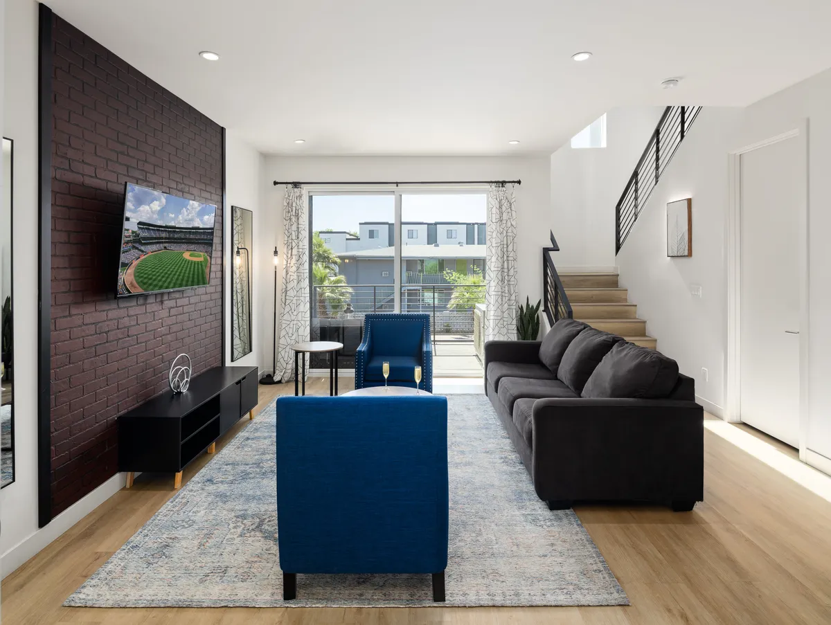 A stylish open-concept living area featuring a dark brown brick accent wall, large window with sheer curtains that filter natural light, blue accent chairs, a cozy sofa, and a wall-mounted Smart TV. The space exudes a contemporary yet cozy vibe, perfect for socializing or relaxing.