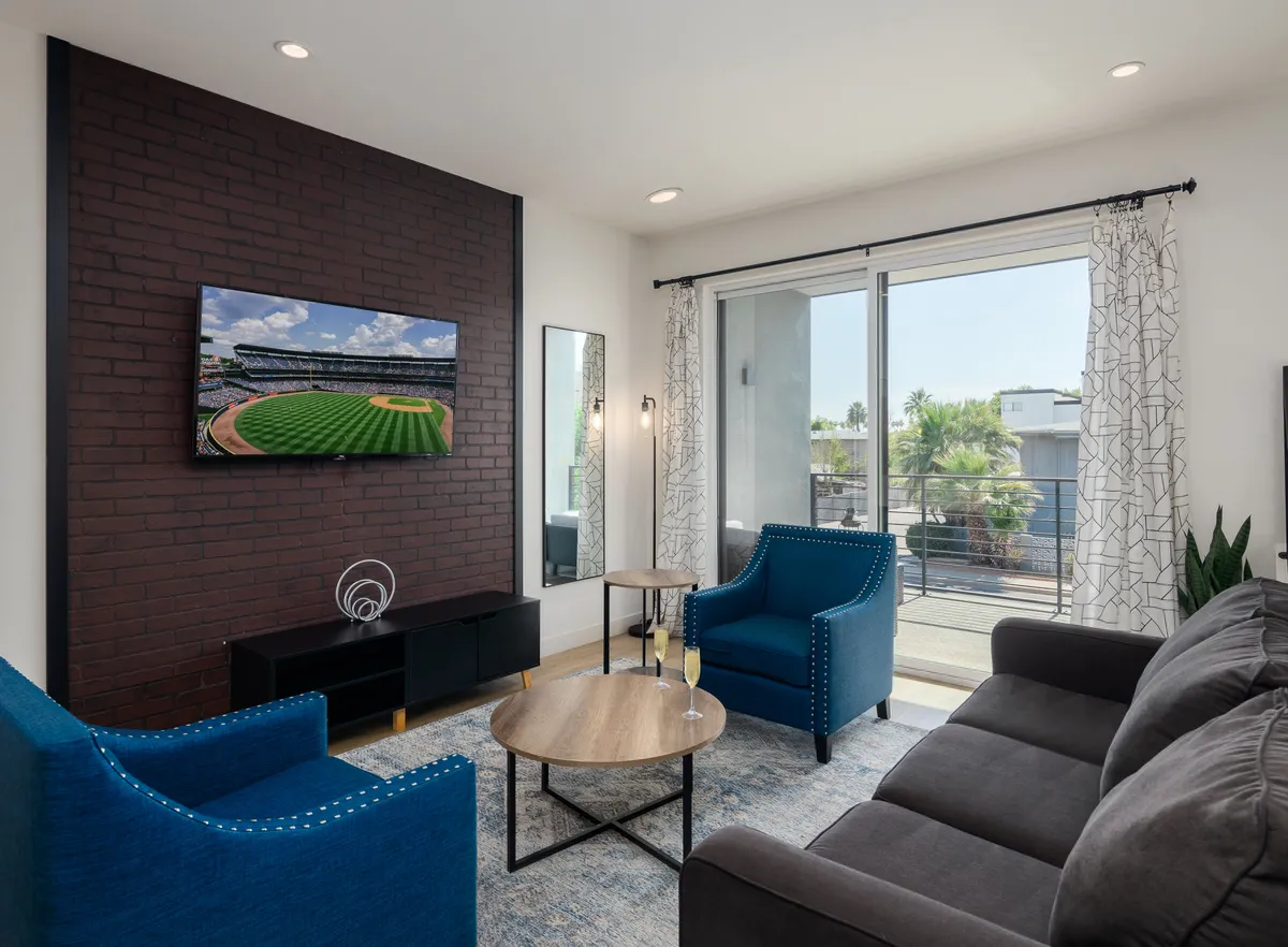 A cozy living area featuring a television displaying a baseball field, comfortable blue chairs, and a wooden coffee table. The setting is casual, ideal for watching sports or relaxing after a busy day.