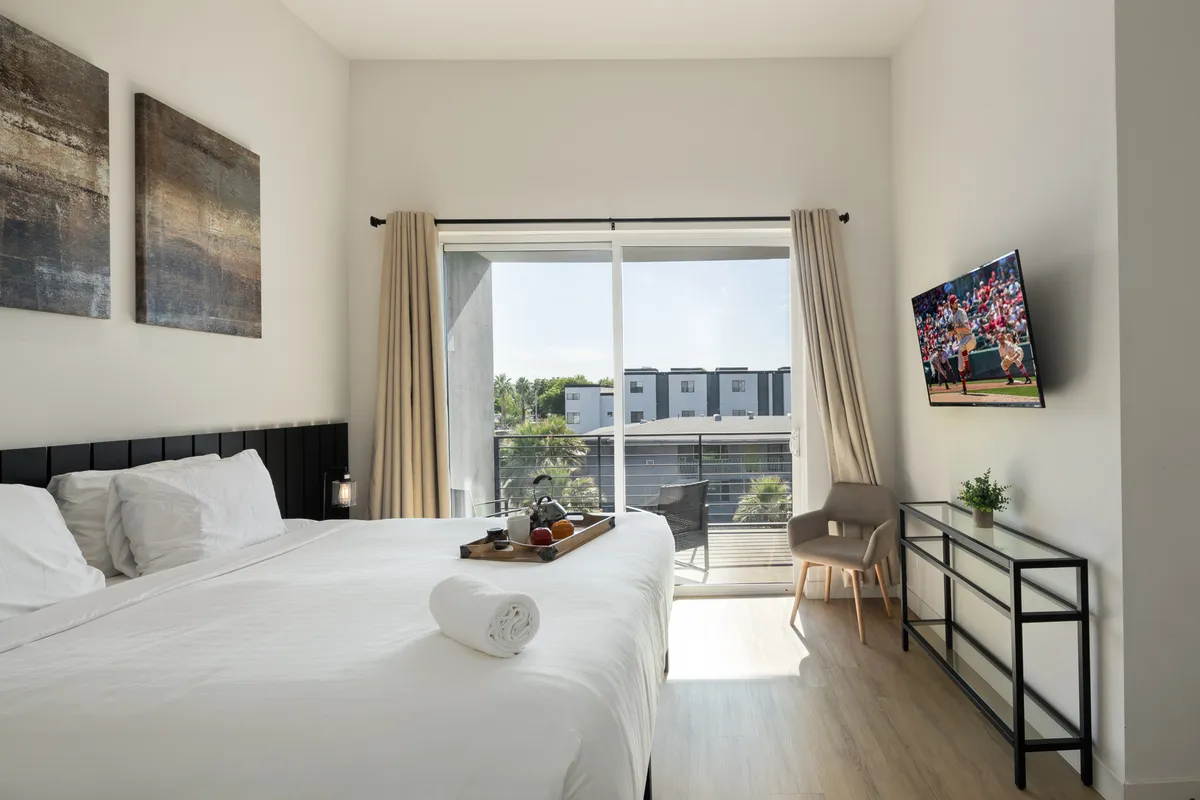 A modern bedroom featuring a plush king bed with crisp linens, a large window letting in morning sunlight, a mounted Smart TV, decorative artwork, and a cozy side table with snacks. The overall mood is serene and welcoming, ideal for restful nights or gentle mornings.