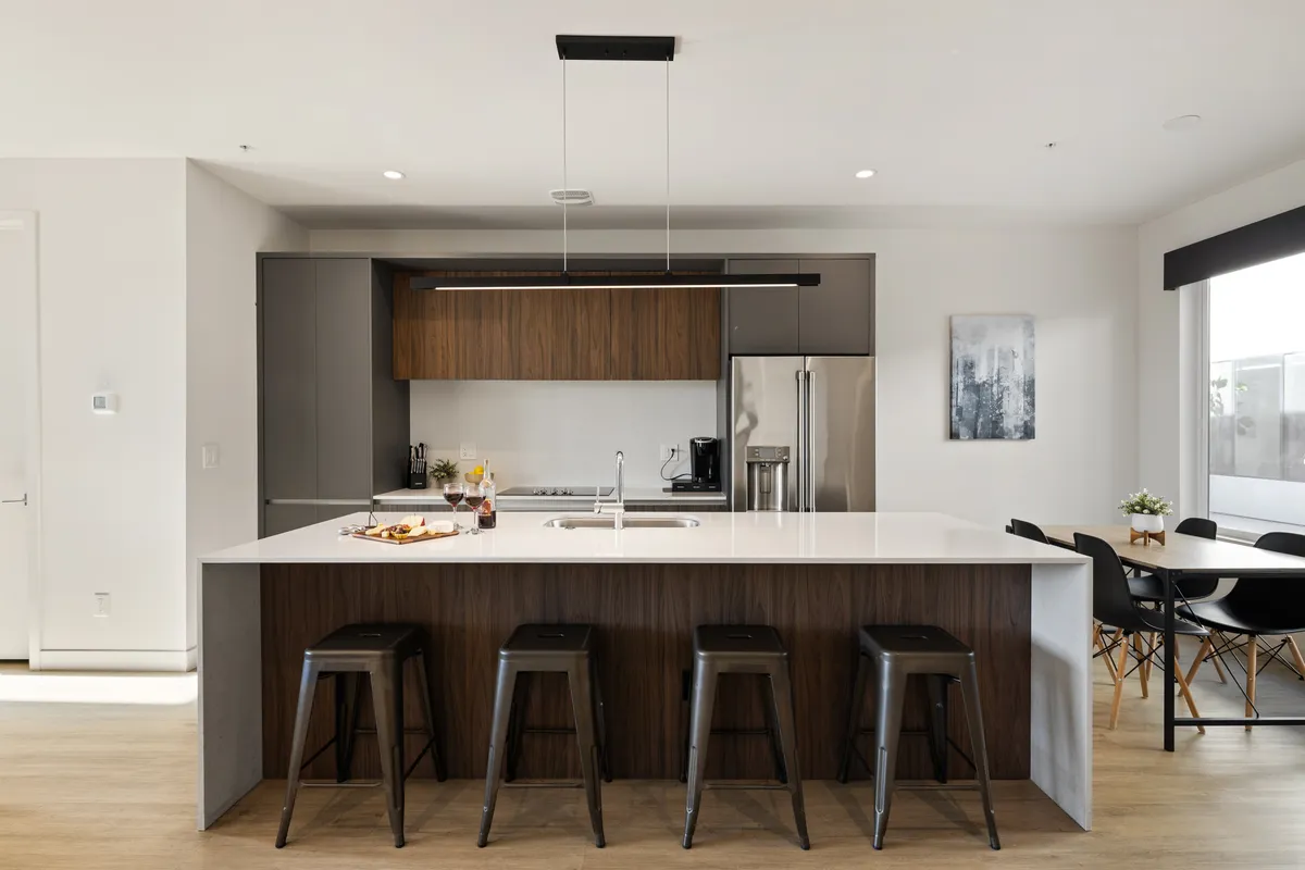 The image showcases a stylish, open-concept kitchen and dining area with a spacious kitchen island surrounded by modern bar stools, stainless steel appliances, tasteful decorative wall art, and an inviting dining table set for gatherings. The space is bathed in natural light, exuding a contemporary yet welcoming atmosphere that invites guests to cook, entertain, and create lasting memories together.