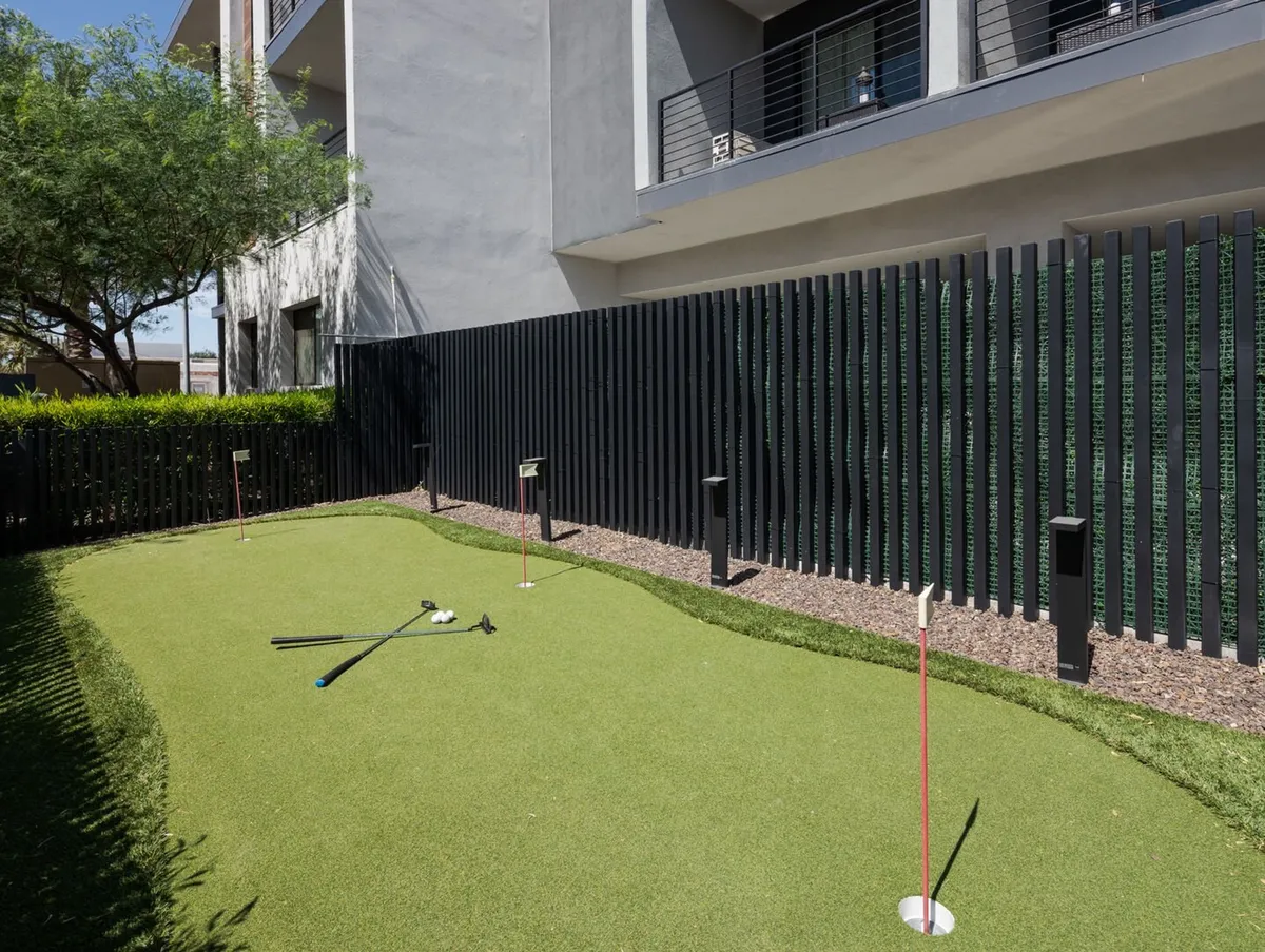 A vibrant outdoor putting green surrounded by neat landscaping, fencing, and the community building facade in the background. The space is designed for casual golf practice or friendly competitions among residents.