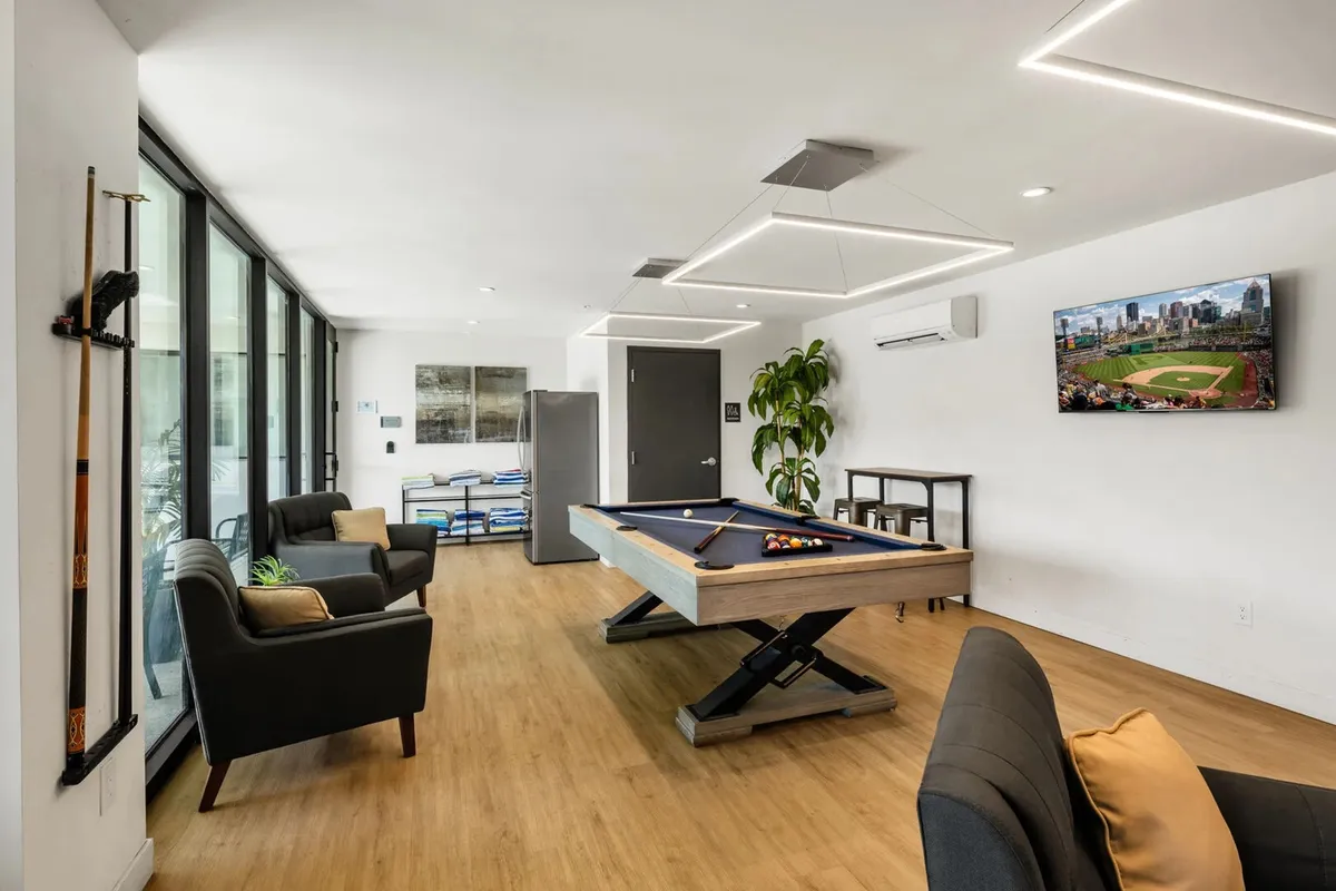 A modern resident clubhouse interior with a pool table, stylish lighting, large windows, and a TV displaying sports. The space is bright and airy, with greenery adding a touch of comfort and community spirit.