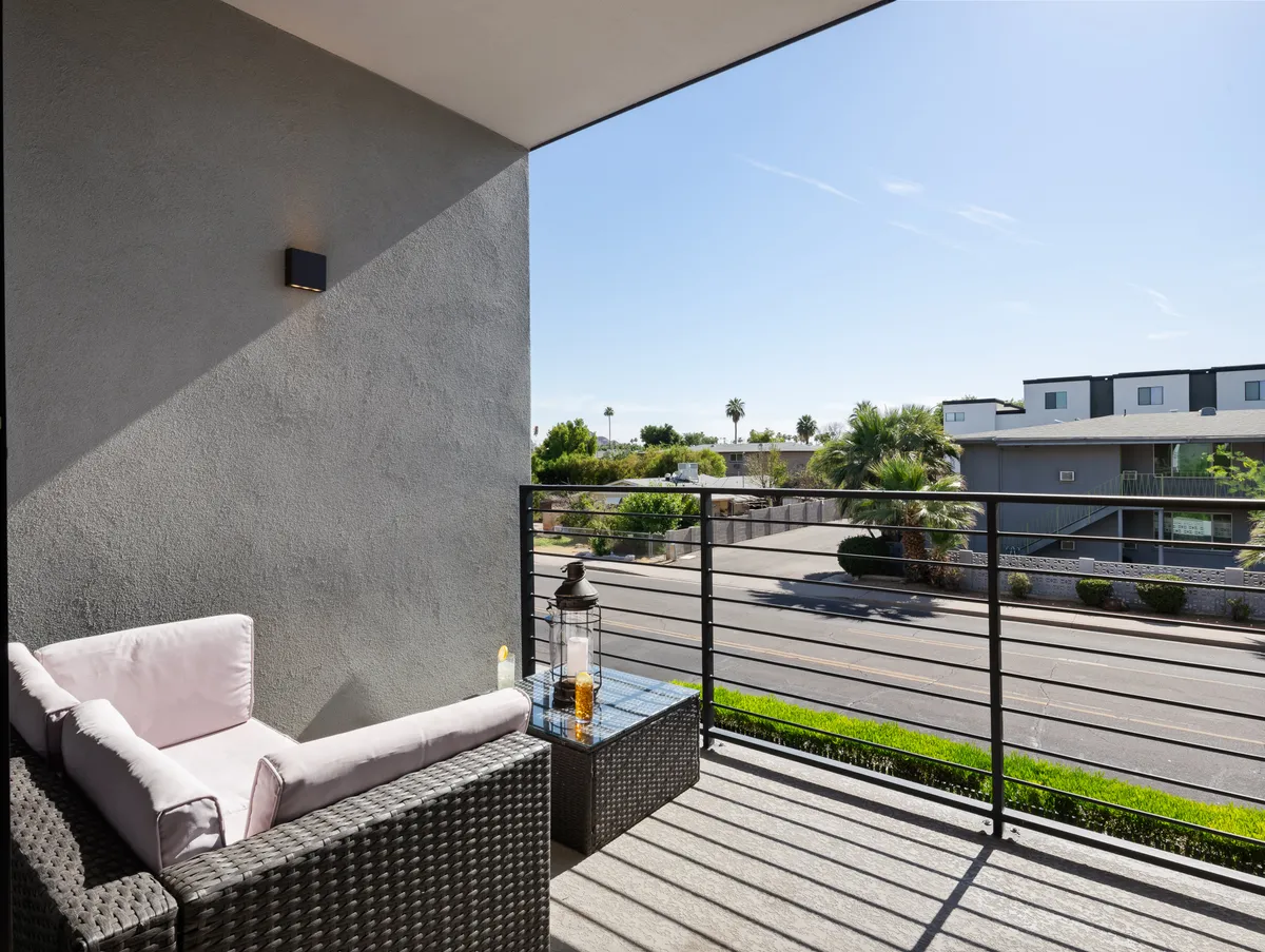 A peaceful exterior setting showing wicker seating and a decorative lantern on a balcony or patio, with a street view beyond. The morning or sunset light creates a calm, inviting mood ideal for enjoying coffee or quiet reflection.