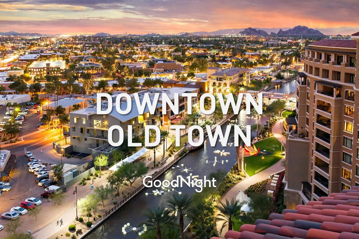 A lively view along the canal in Old Town Scottsdale, with illuminated buildings, palm trees, and mountains in the background. People are seen strolling, dining at outdoor cafes, and exploring local shops as the evening lights bring the area to life.