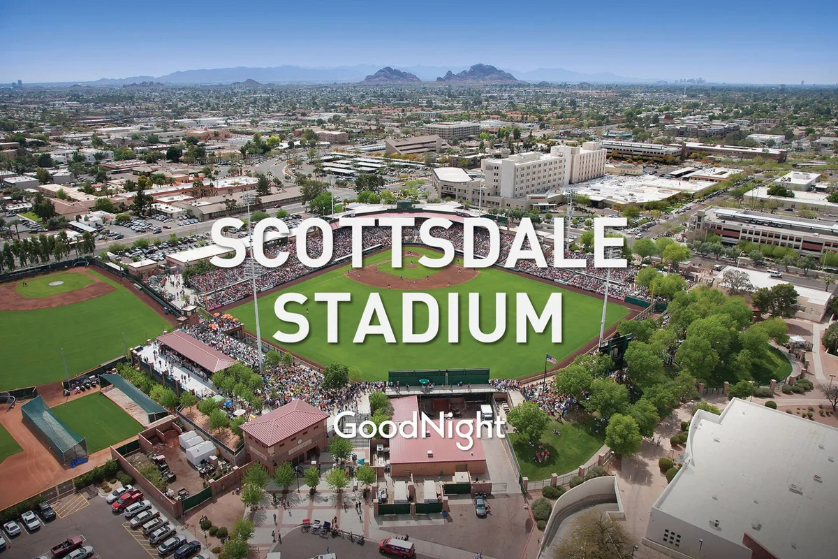 A vibrant baseball field buzzes with energy as fans gather in the spectator stands. Surrounded by city buildings and framed by scenic mountains, the scene conveys the excitement of a live sporting event and the communal spirit of cheering for a favorite team.