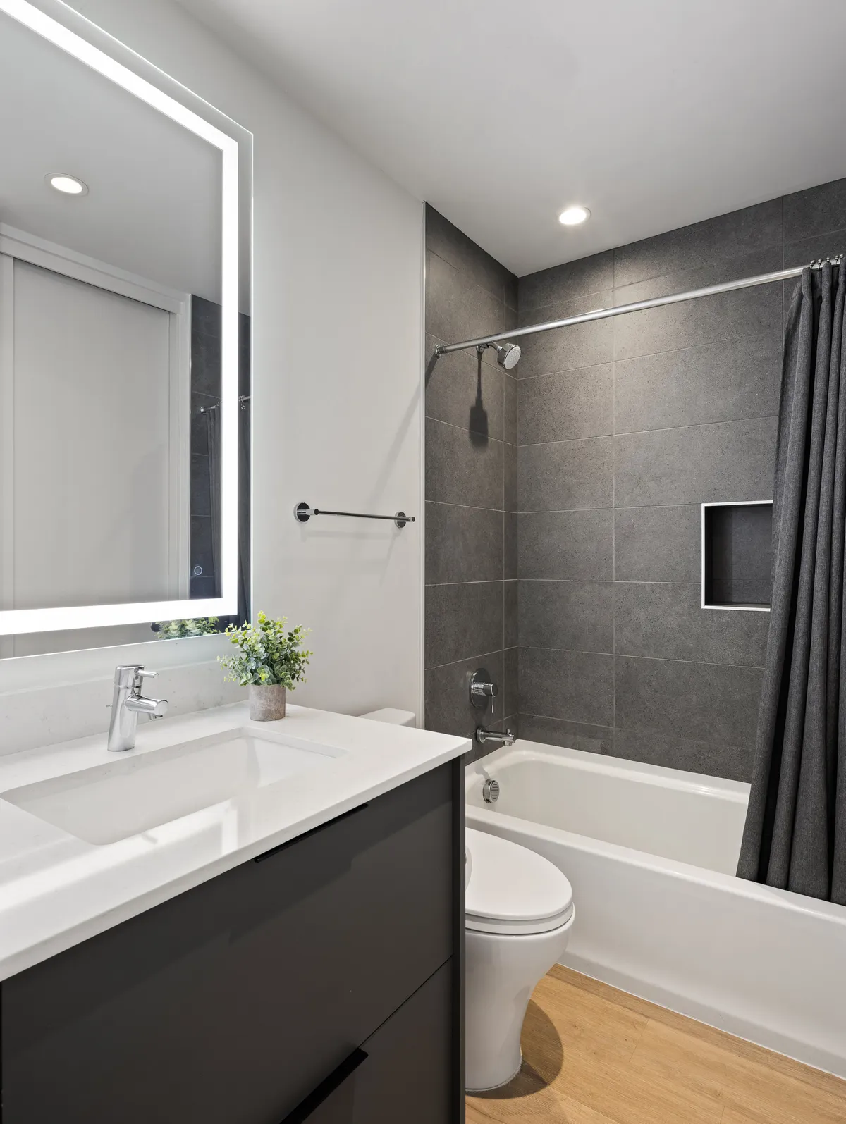 A modern bathroom with a stylish mirror featuring LED lighting, a sleek sink, and a bathtub with a shower curtain. A small potted plant adds a touch of homeliness and serenity.