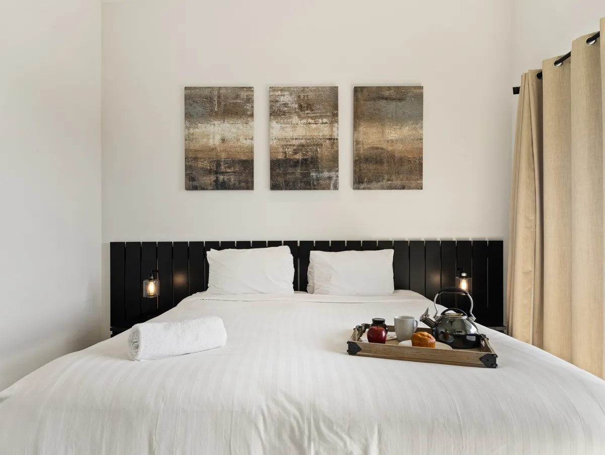 A serene bedroom with white linens, elegant artwork above the bed, and a breakfast tray set for a leisurely morning. The soft lighting and cozy atmosphere create a sense of intimacy and comfort, reflecting a peaceful retreat.