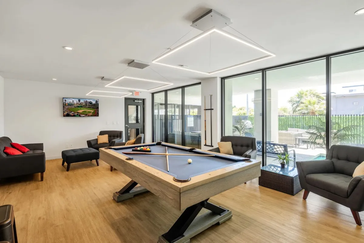 A spacious and bright clubhouse interior featuring a pool table as the focal point, surrounded by comfortable sofas, a large television, and expansive windows letting in natural light. The setup invites both casual billiards games and social gatherings.